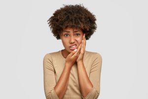 Woman in a brown shirt holding 2 hands to her jaw in pain in front of a white background